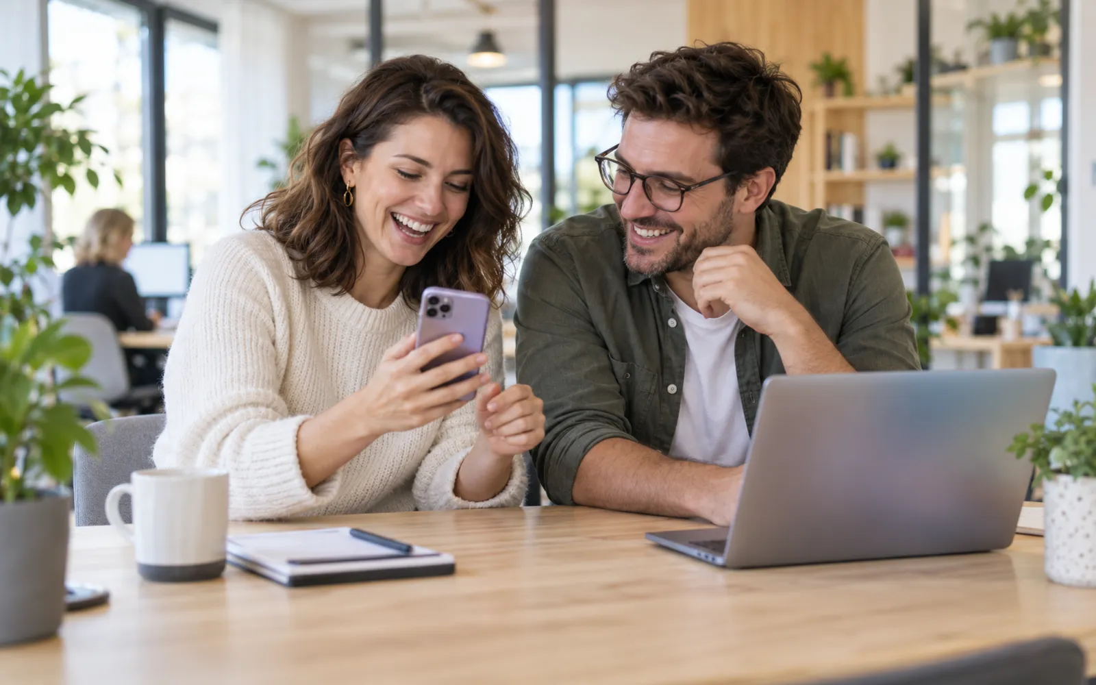 Photo: a woman and a man sit at a wooden table in a bright office; she shows him something on her smartphone and both laugh