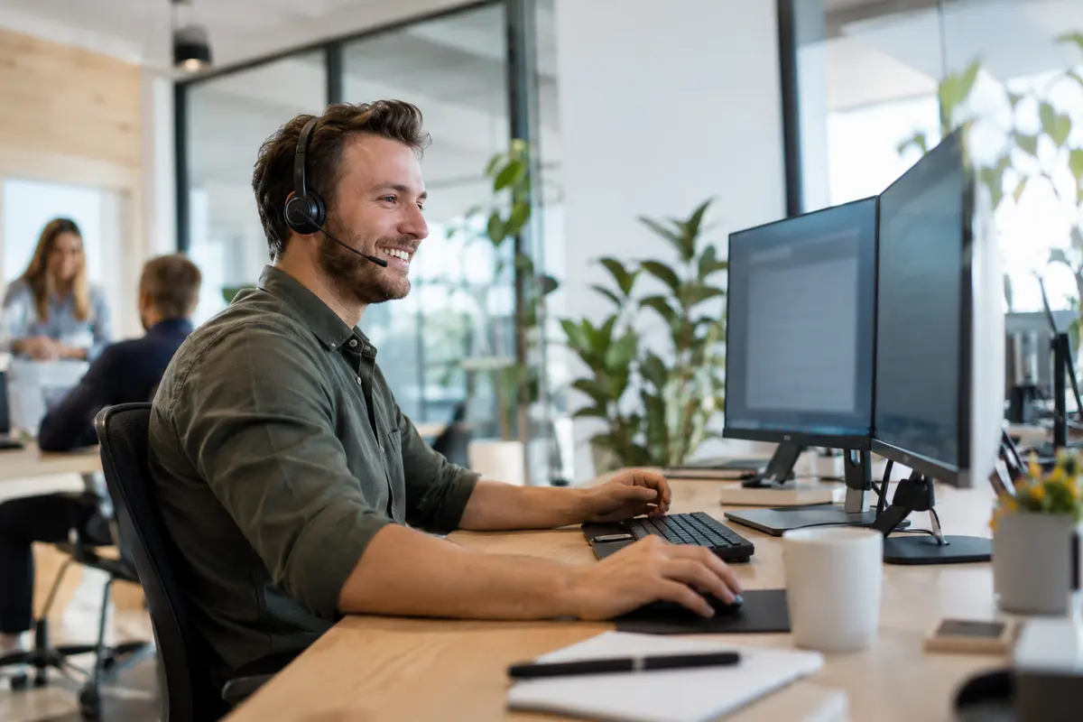 Photo: a man with a headset sits smiling at a desk with two monitors in a bright open-plan office