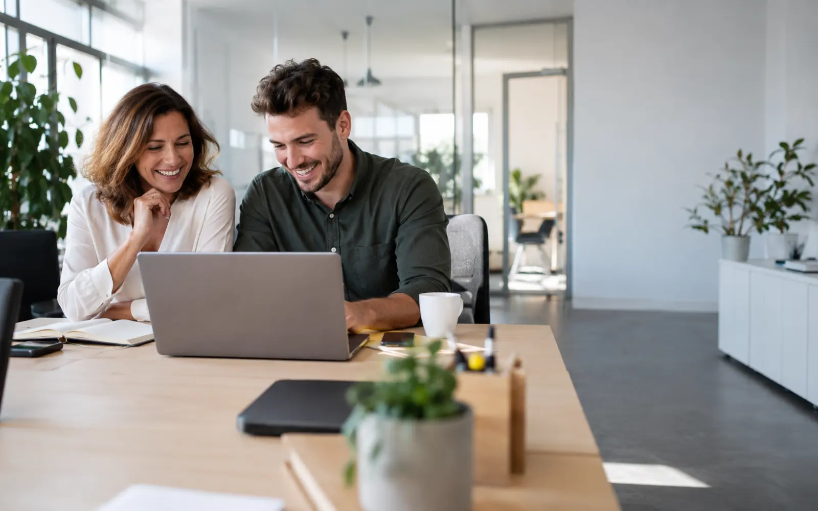Photo: a woman and a man sit at a wooden table in a bright office, look at a laptop together and laugh